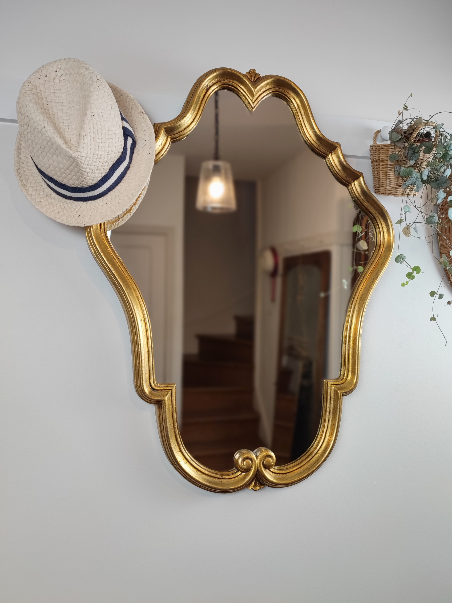 Grand miroir baroque "Angèle" mouluré finition doré. Un vrai miroir de conte de fée à installer dans une entrée. Elégant, chic et sobre à la fois. Chapeau de paille et plante verte dans un panier accroché à une patère murale.