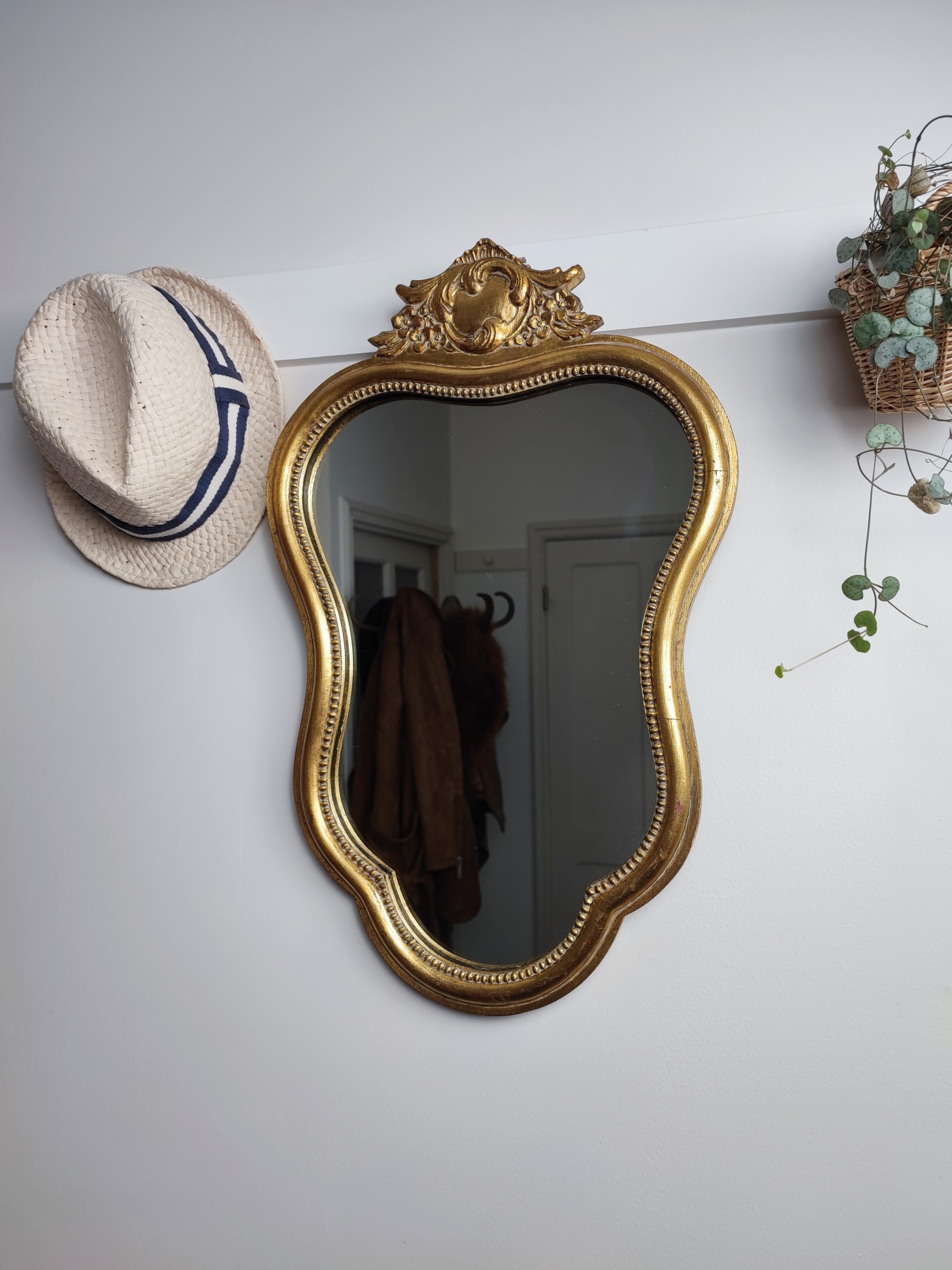 Miroir baroque "Yvette" en bois mouluré, finition doré patiné avec un joli fronton sculpté à son sommet. Inscription Noël 1968 au dos du miroir. Chapeau de paille. Plante verte.