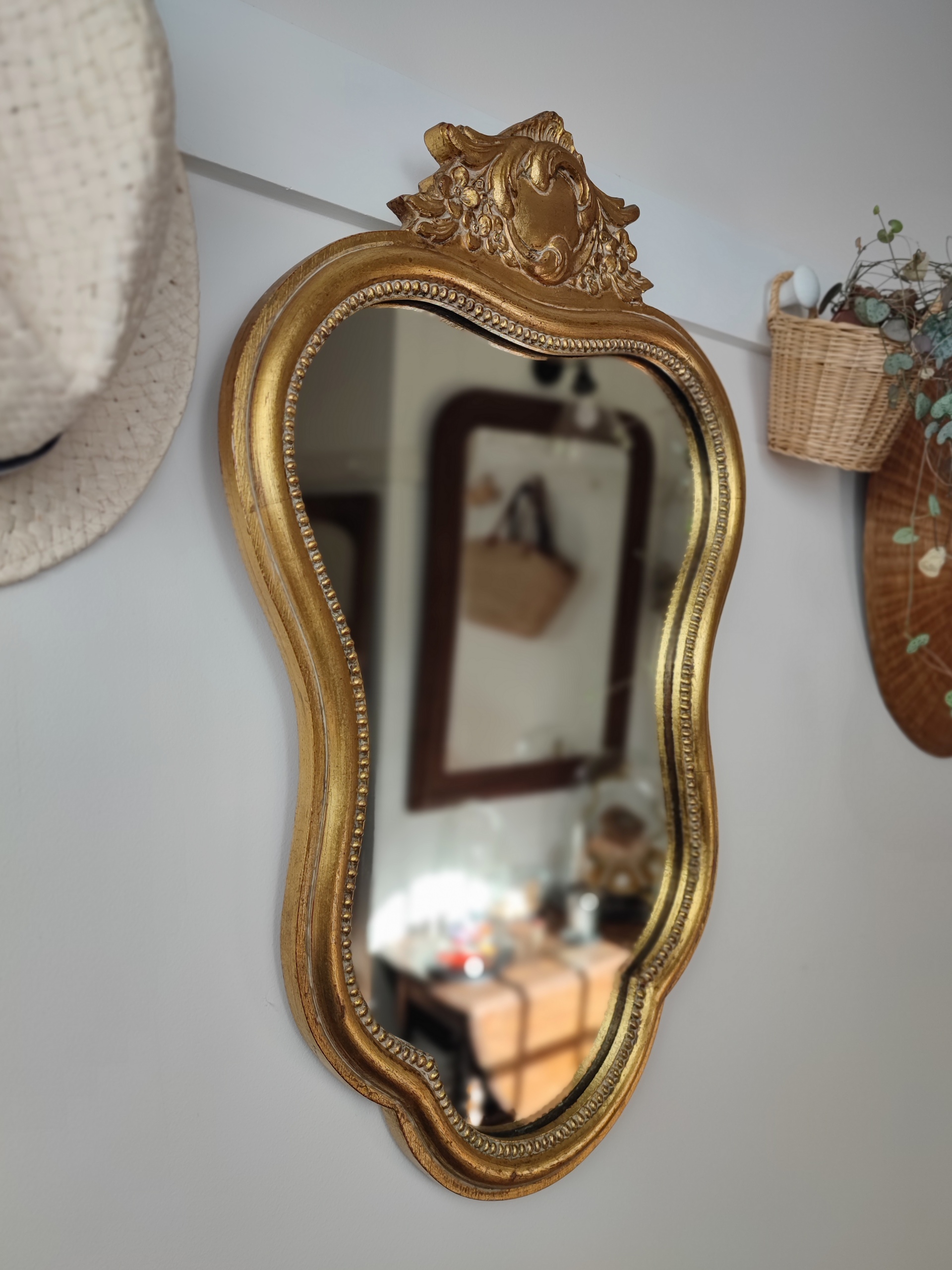 Miroir baroque "Yvette" en bois mouluré, finition doré patiné avec un joli fronton sculpté à son sommet. Inscription Noël 1968 au dos du miroir. Chapeau de paille. Plante verte.
